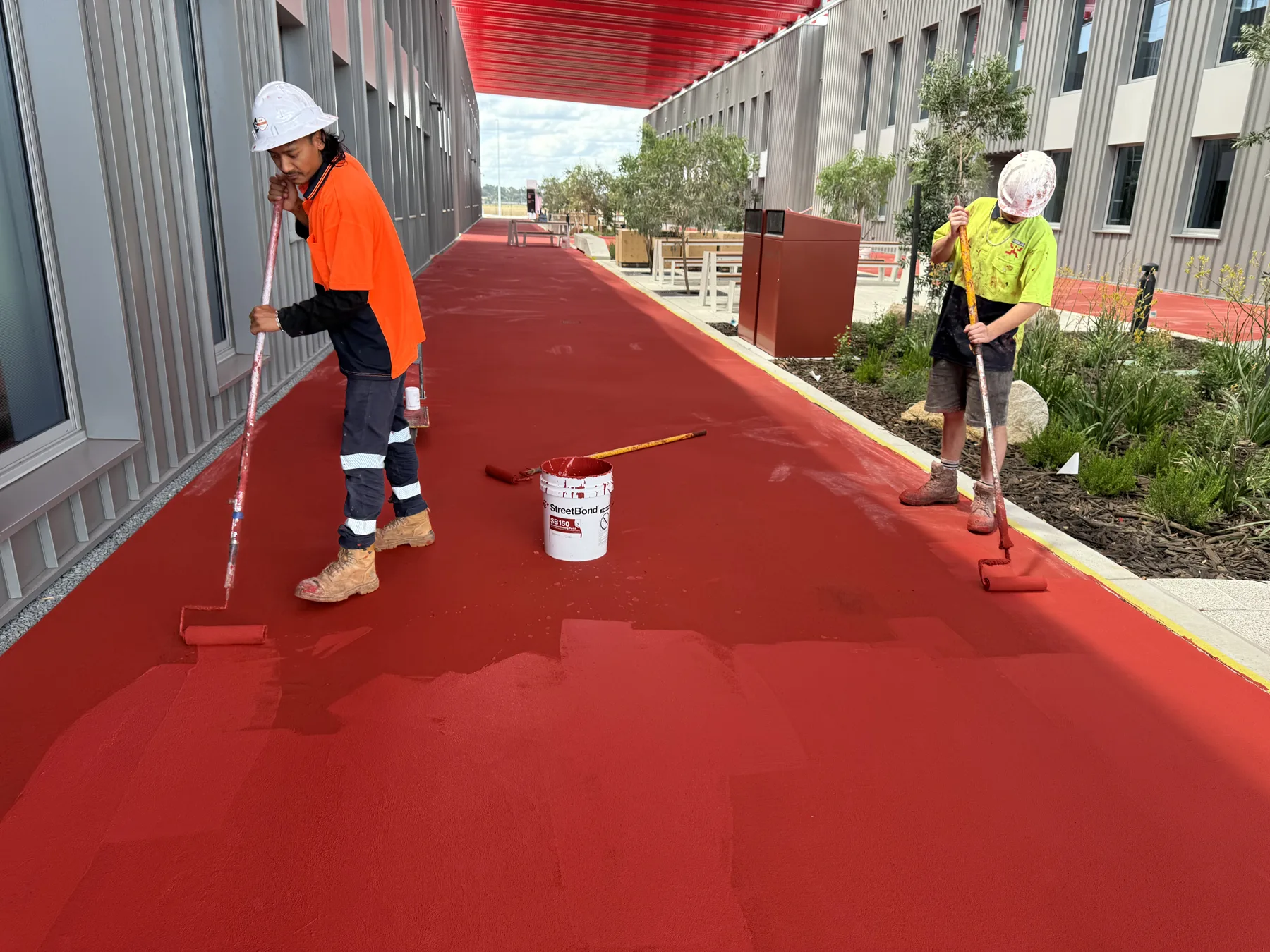 Two applicators rolling red StreetBond SB150 coating onto a concrete walkway between commercial buildings